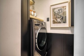 A laundry room with a front-loading washer and dryer, a shelf with jars, and a framed abstract painting on the wall.