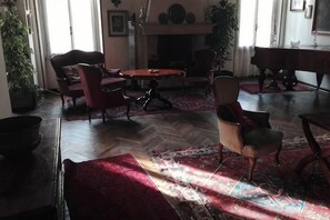 Ein Zimmer mit Kamin, Klavier und Esstisch mit Stühlen.