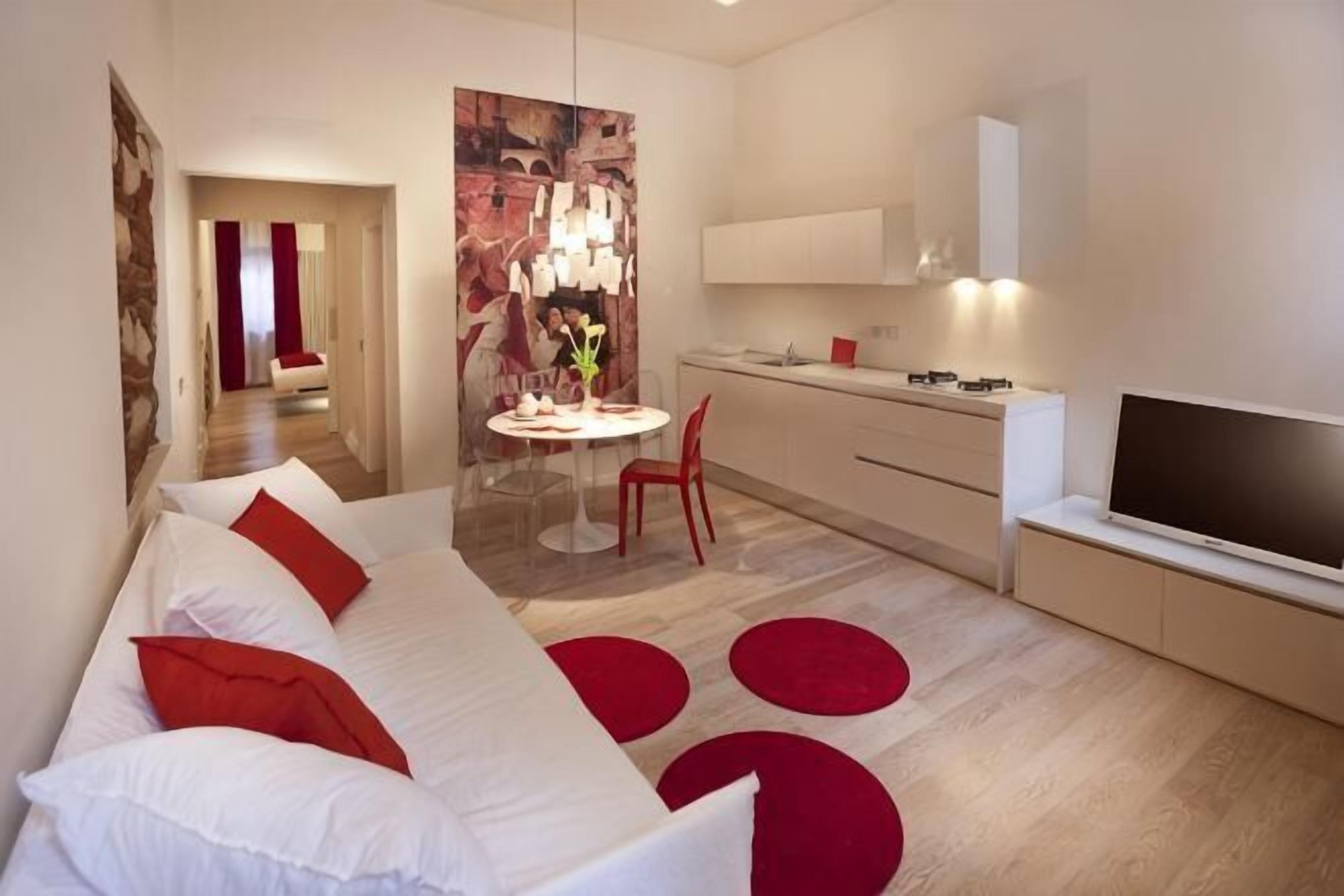 A modern hotel room with a sofa, a dining table, a TV, and a kitchen area.