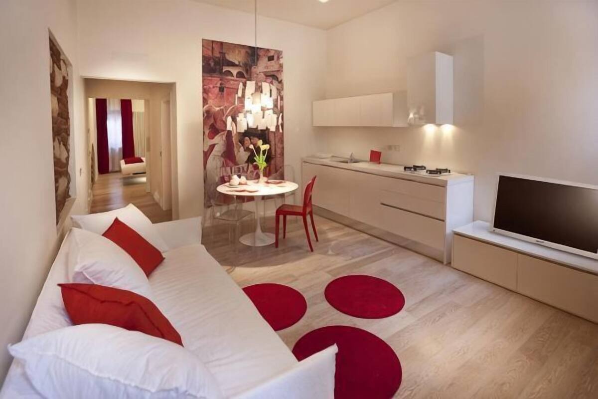 A modern hotel room with a sofa, a dining table, a TV, and a kitchen area.