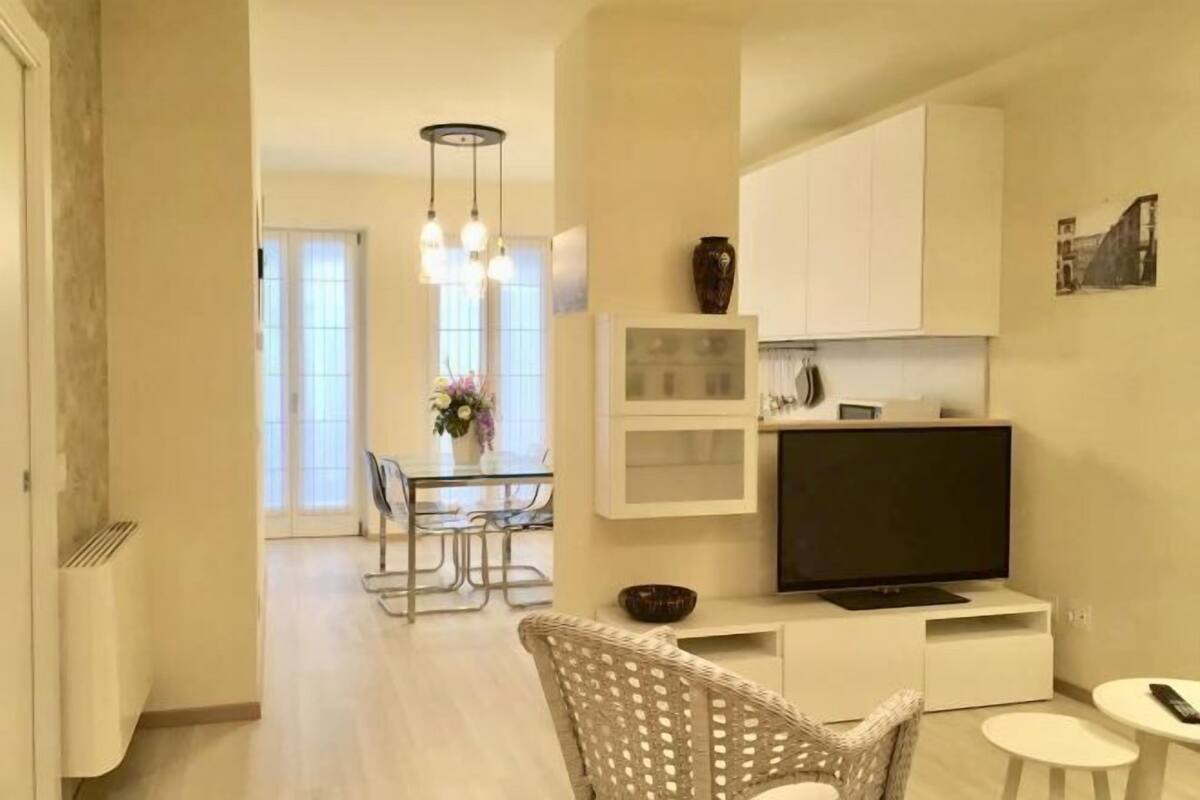 A modern living room with a flat-screen TV, a dining area with a glass table, and a kitchen with white cabinets.