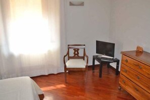 A room with a wooden floor, a white bed, a wooden chair, a TV on a stand, and a wooden dresser. - Truly Verona (Verona)