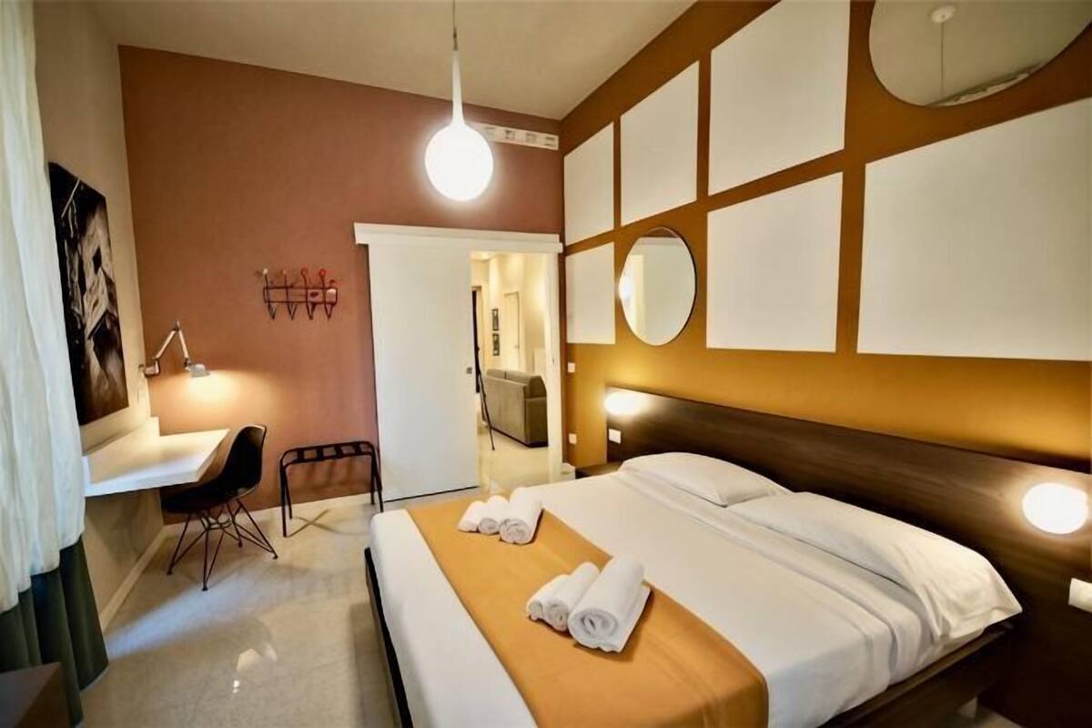 A hotel room with a bed, a desk, a chair, a mirror, and a wall-mounted light fixture.