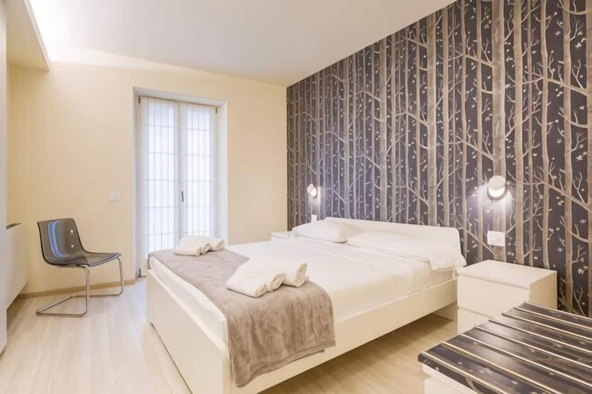 A bedroom with a bed, bedside table, chair, and a window with blinds.