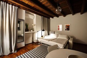 A bedroom with a bed, a bedside table, a chair, a radiator, a mirror, and a window with curtains. - Truly Verona (Verona)