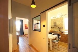 Un couloir mène à une salle à manger avec une table et des chaises, une cuisine équipée d’une cuisinière, et une porte avec une poignée.