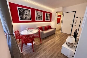 A modern living room with a red accent wall, a grey sofa, a red dining table with chairs, and a white coffee table. - Truly Verona (Verona)