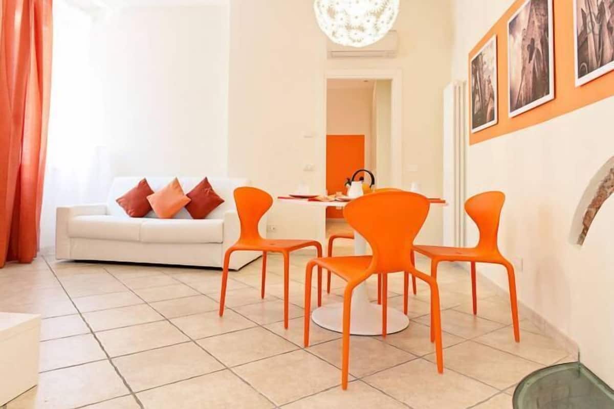 A modern living room with a white sofa, orange chairs, and a dining table.