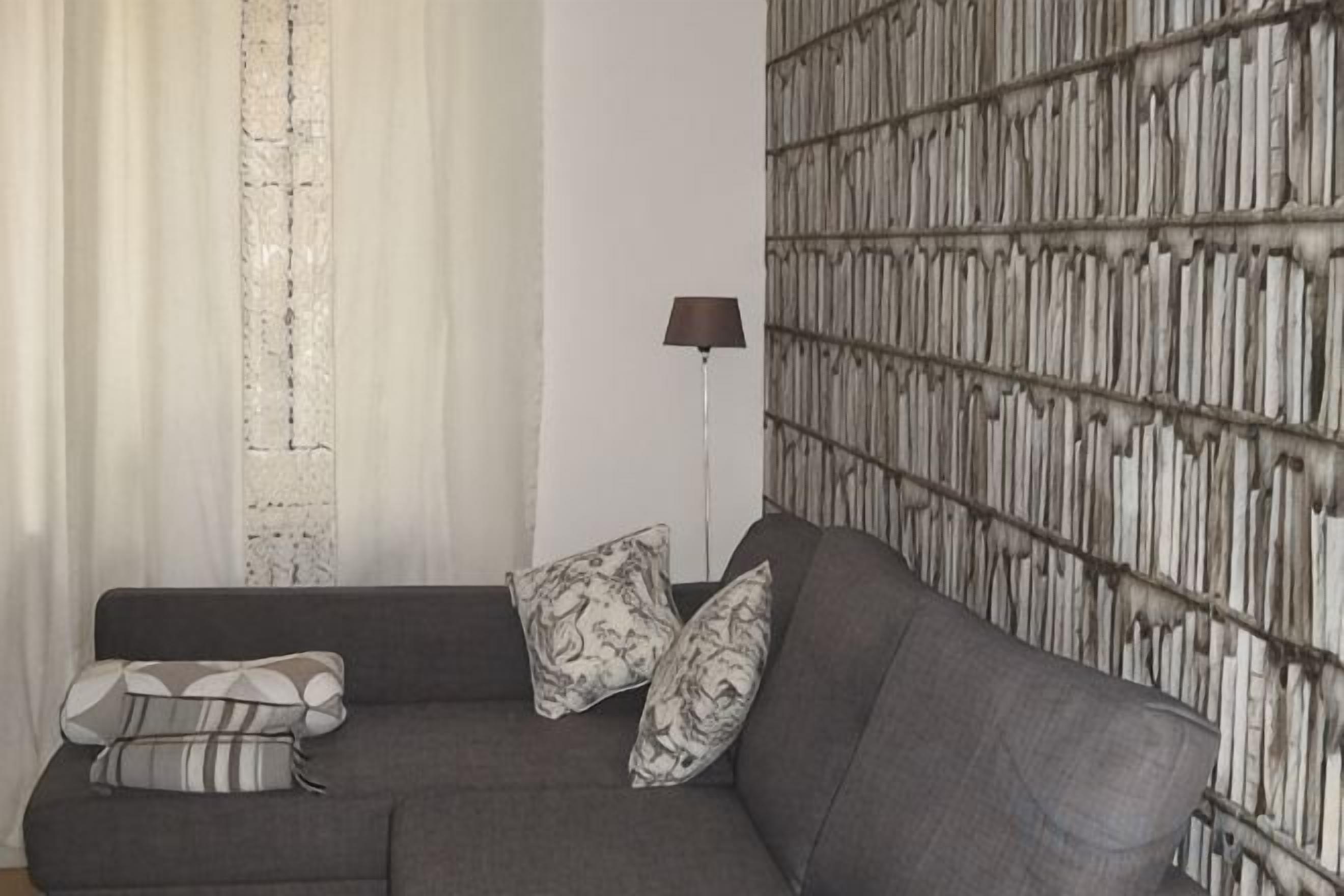 Ein graues Sofa mit gemusterten Kissen, eine Stehlampe und eine Wand mit vertikalem Bücherregal-Muster.