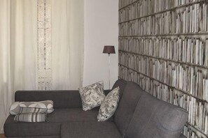 A grey sofa with patterned cushions, a floor lamp, and a wall with a vertical book pattern. - Truly Verona (Verona)