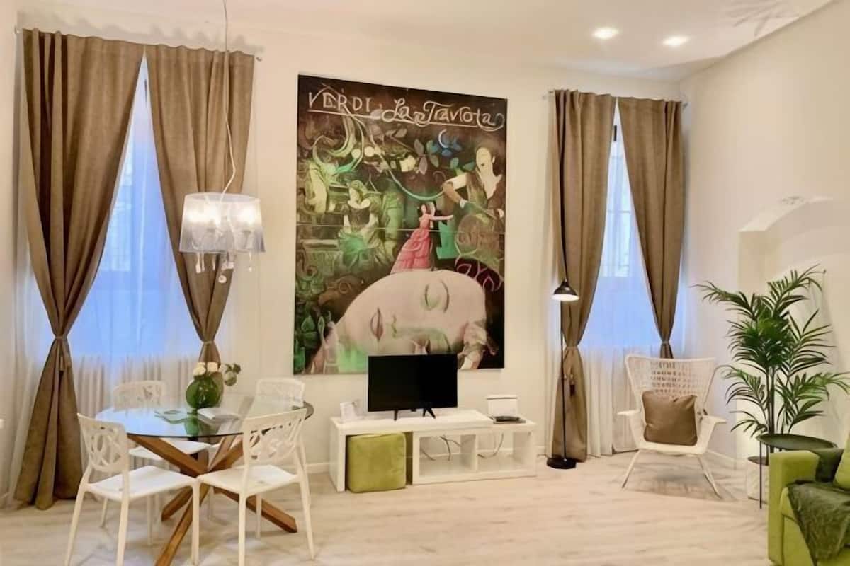 A modern living room with a flat-screen TV, white furniture, a large abstract painting, and a potted plant.