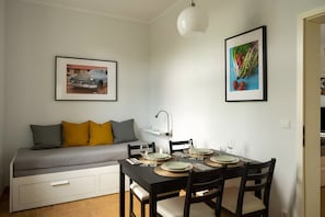 Un coin repas avec une table dressée pour quatre personnes, un canapé avec des coussins, un tableau encadré représentant une voiture et un autre tableau encadré représentant des légumes.