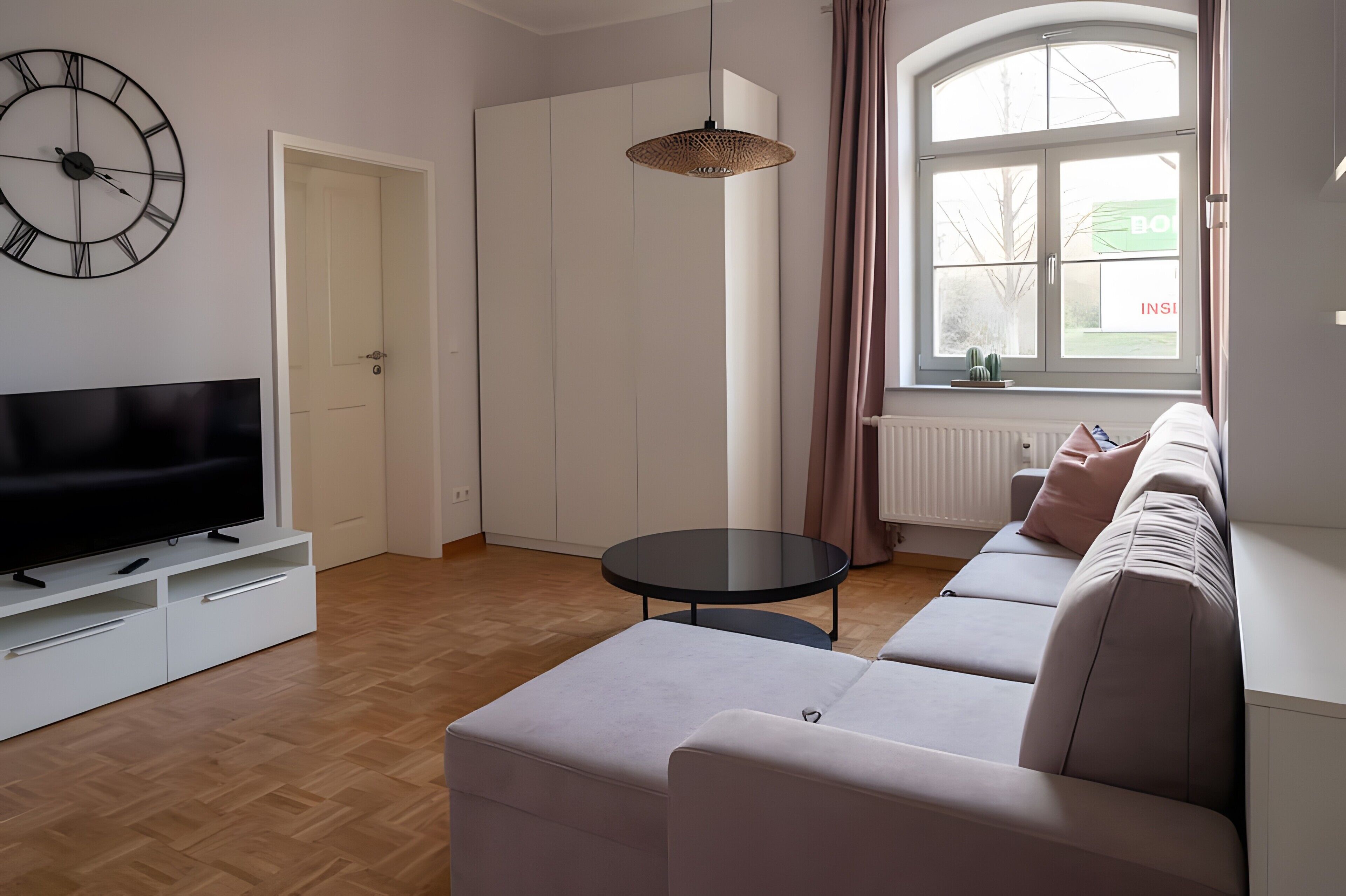 Un salon moderne avec un téléviseur à écran plat, une table ronde, un ensemble de canapés, une horloge et une fenêtre avec des rideaux.