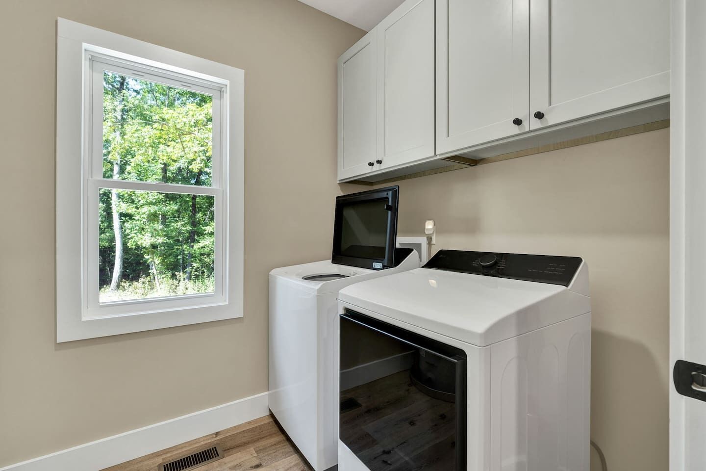 A laundry room with a washer and dryer, a microwave, and a window with a view of trees.