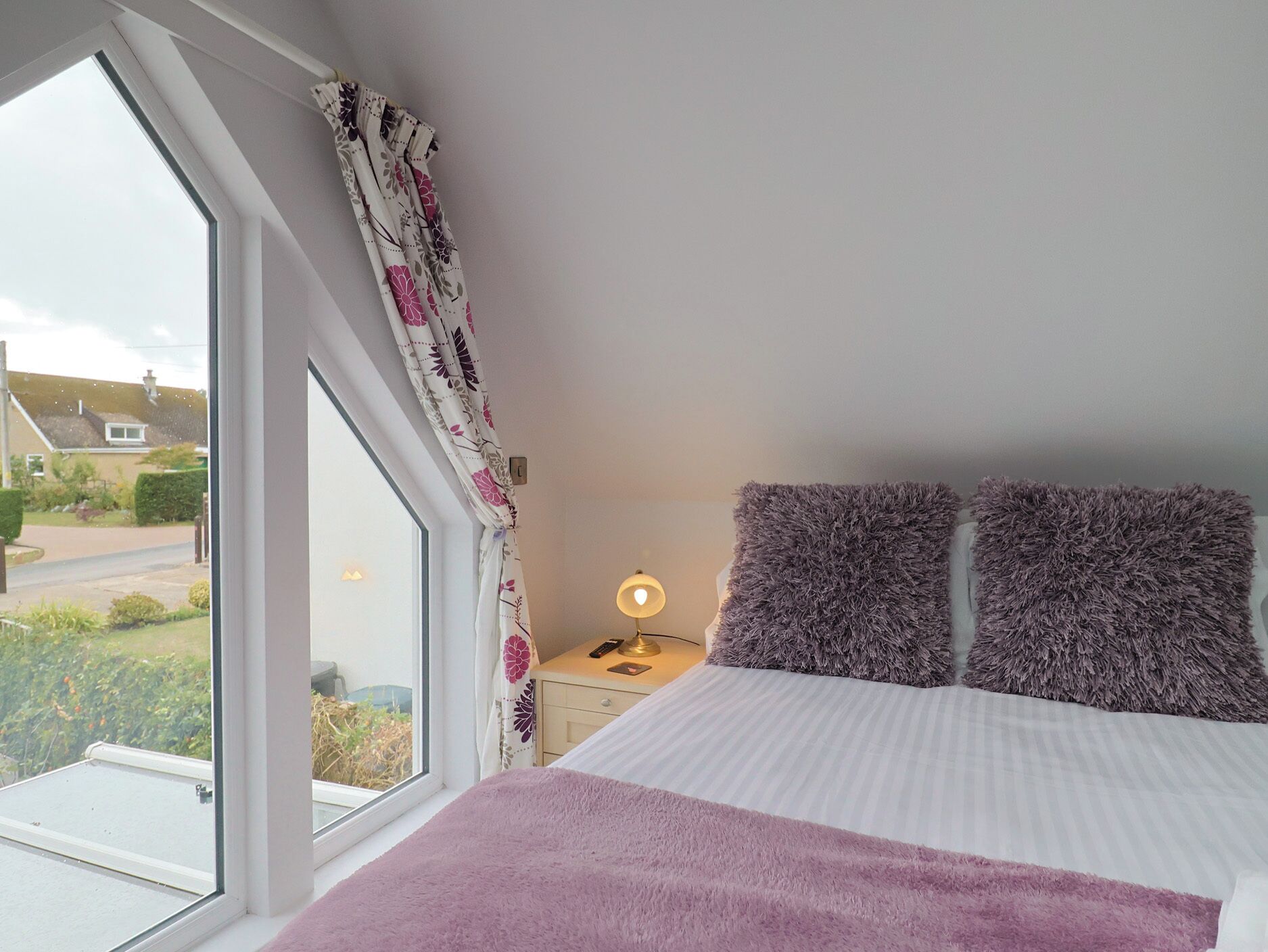 A bedroom with a large window, a bed with a purple blanket, and a nightstand with a lamp.