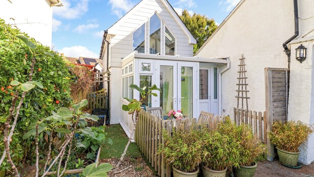 A small, white, glass-enclosed structure with a sloped roof, surrounded by potted plants and a wooden fence. - Cherry Pip (Bridport)
