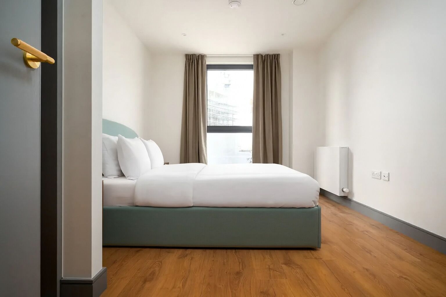 Een moderne slaapkamer met een bed, een raam met gordijnen, een radiator en een deur met gouden handvat.
