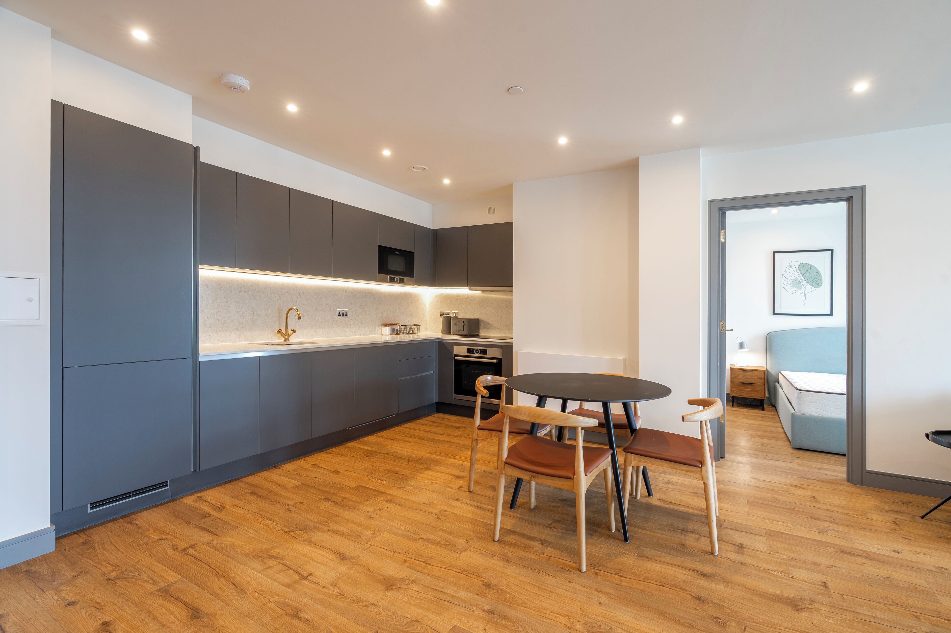 A modern kitchen with grey cabinets, a wooden dining table, and a blue sofa in the adjacent room.