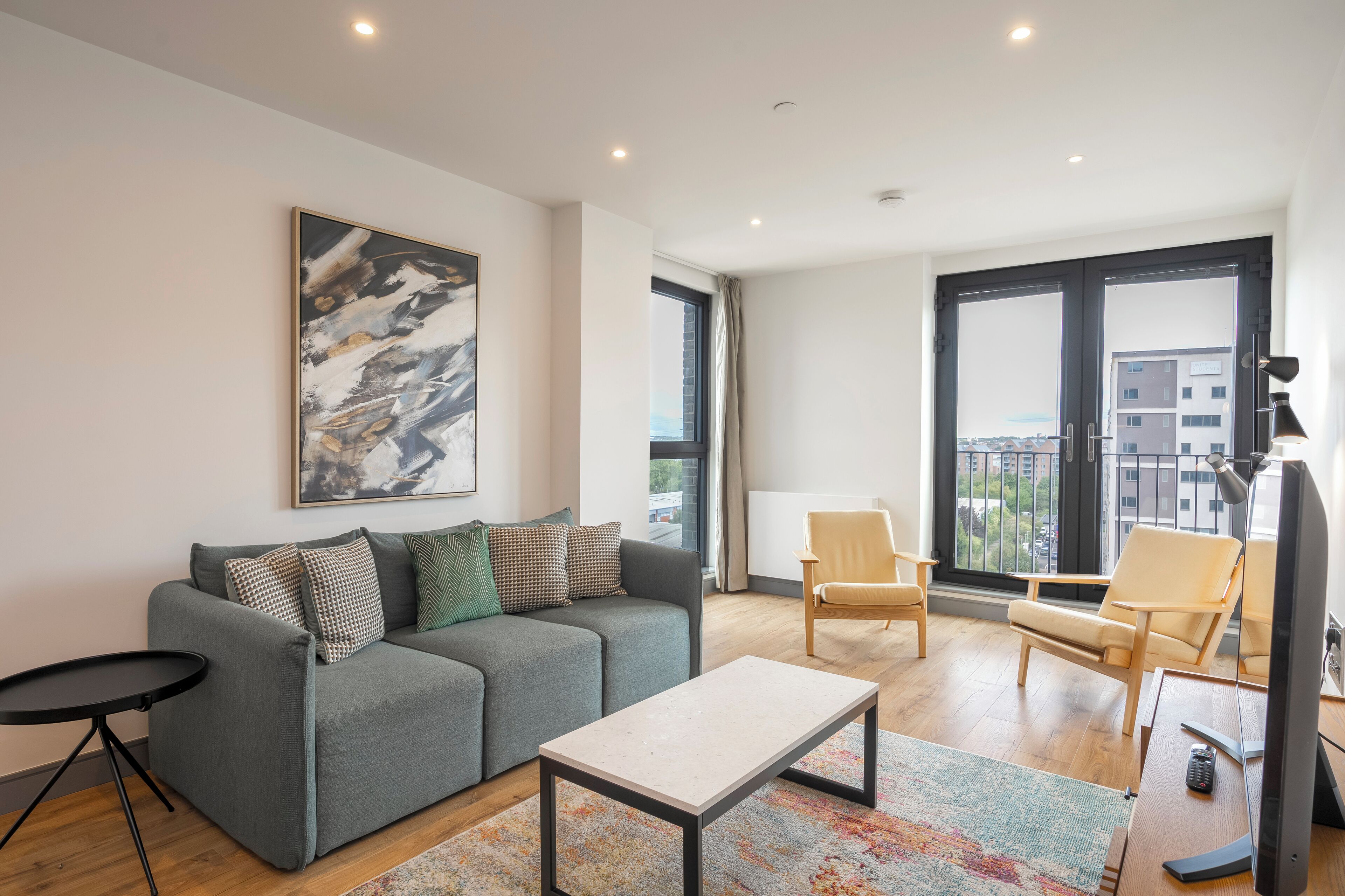 A modern living room with a grey sofa, a coffee table, and a TV. There are two chairs and a large window with a view of buildings.