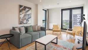 Una sala moderna con un sofá gris, una mesa de centro y un televisor. Hay dos sillones y un ventanal con vistas a edificios.