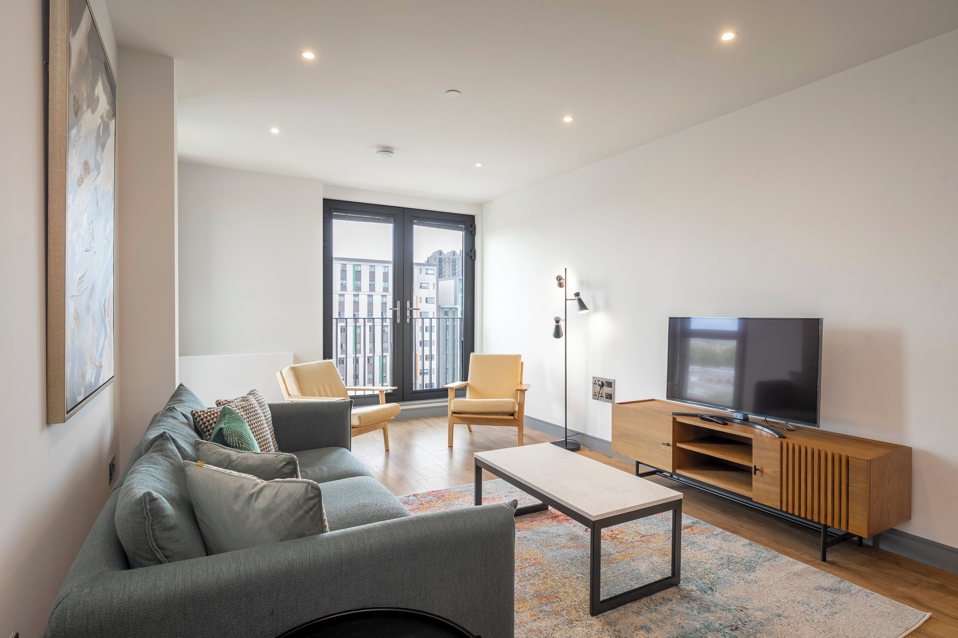 Una sala de estar moderna con un sofá gris, una mesa de centro, un televisor sobre un mueble de madera y un ventanal con vistas a la ciudad.