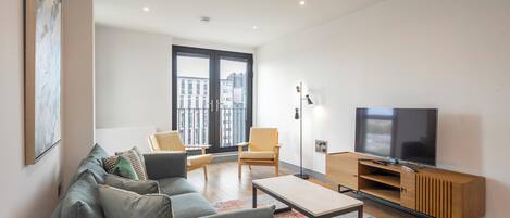 Una sala de estar moderna con un sofá gris, una mesa de centro, un televisor sobre un mueble de madera y un ventanal con vistas a la ciudad.