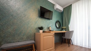 A room with a teal textured wall, a flat-screen TV, a wooden bench, a small cabinet, a desk with a chair, and a window with sheer curtains. - Thaya Rooms (Mangalia)
