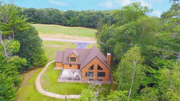 A two-story log cabin with a wrap-around porch, surrounded by dense forest.