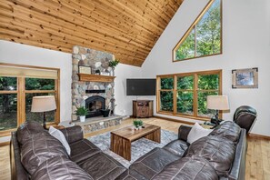 A living room with a stone fireplace, leather sofas, a flat-screen TV, and large windows with a view of greenery. - Stepaway Vacation Rentals- Stay-A-While (Eagle River)