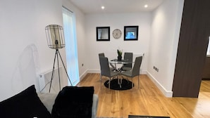 A modern living room with a grey sofa, a coffee table, and a dining area with a small table and chairs. There is a floor lamp and a clock on the wall.
