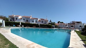 A rectangular swimming pool with a concrete deck, surrounded by a well-maintained lawn and a row of white buildings with red-tiled roofs. - Charming cottage in a quiet environment (Nerja)