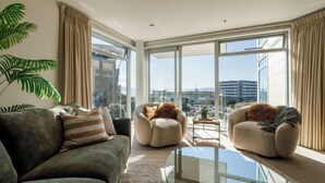 A modern living room with a sofa, two armchairs, a glass coffee table, and large windows offering a city view.