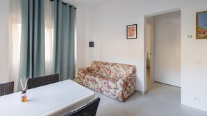 Una habitación con un sofá blanco, una mesa de comedor y una silla. Hay un cuadro en la pared y una puerta que da a otra habitación.