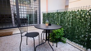 Un pequeño patio al aire libre con una mesa redonda y dos sillas, una planta en maceta sobre la mesa y una valla metálica con follaje al fondo.