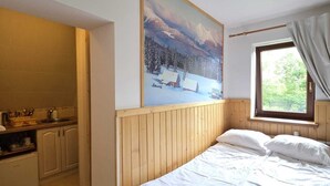 Ein Schlafzimmer mit einem hölzernen Bett, einem Gemälde einer verschneiten Landschaft, einem Fenster mit Vorhängen und einer Küchenzeile im angrenzenden Raum.