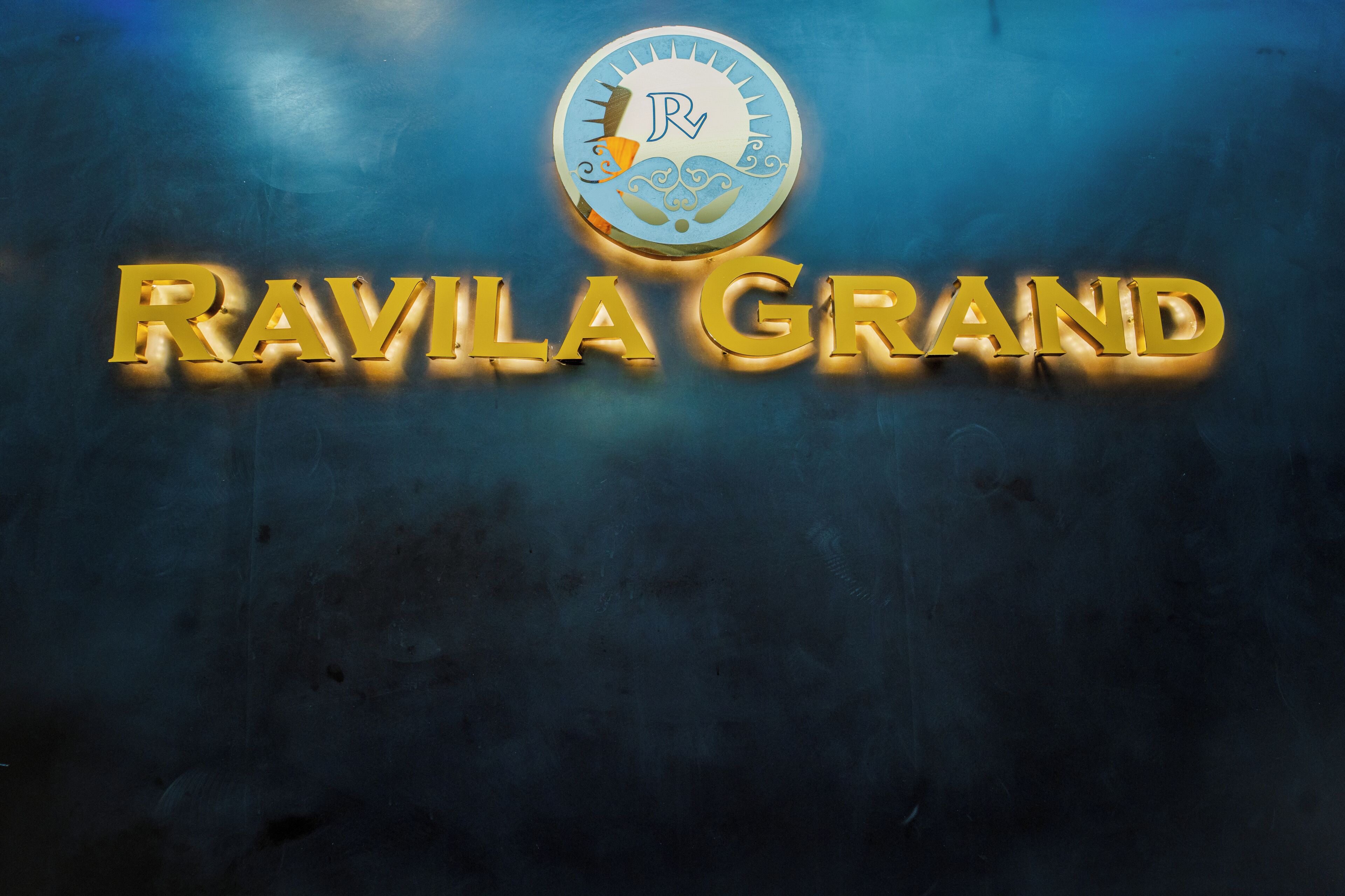 Ein Logo mit dem Text 'RAVILA GRAND' und einem kreisförmigen Emblem mit Sonne und Wellen.