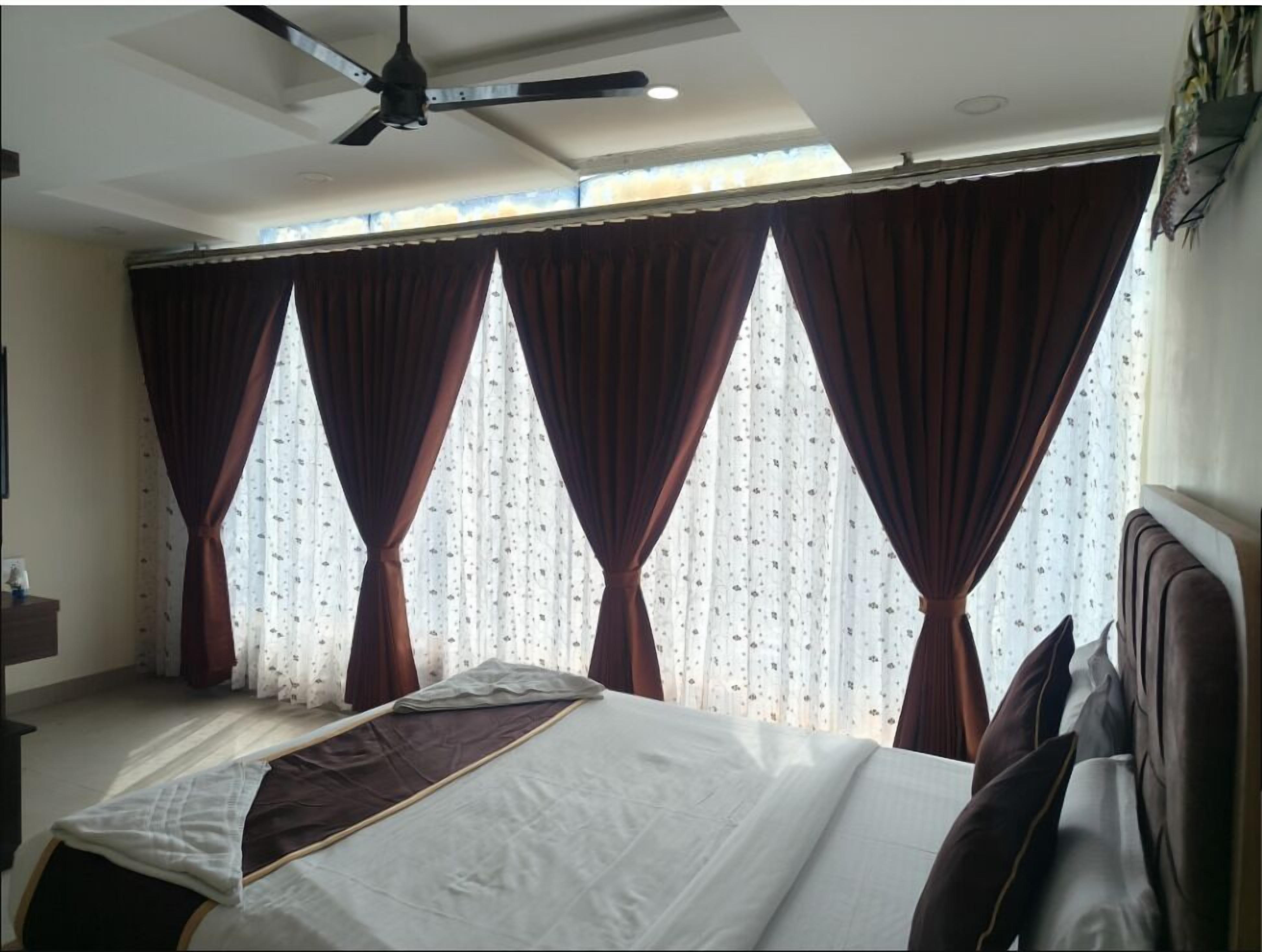 Habitación de hotel con cama, ventilador de techo y ventanas amplias con cortinas.