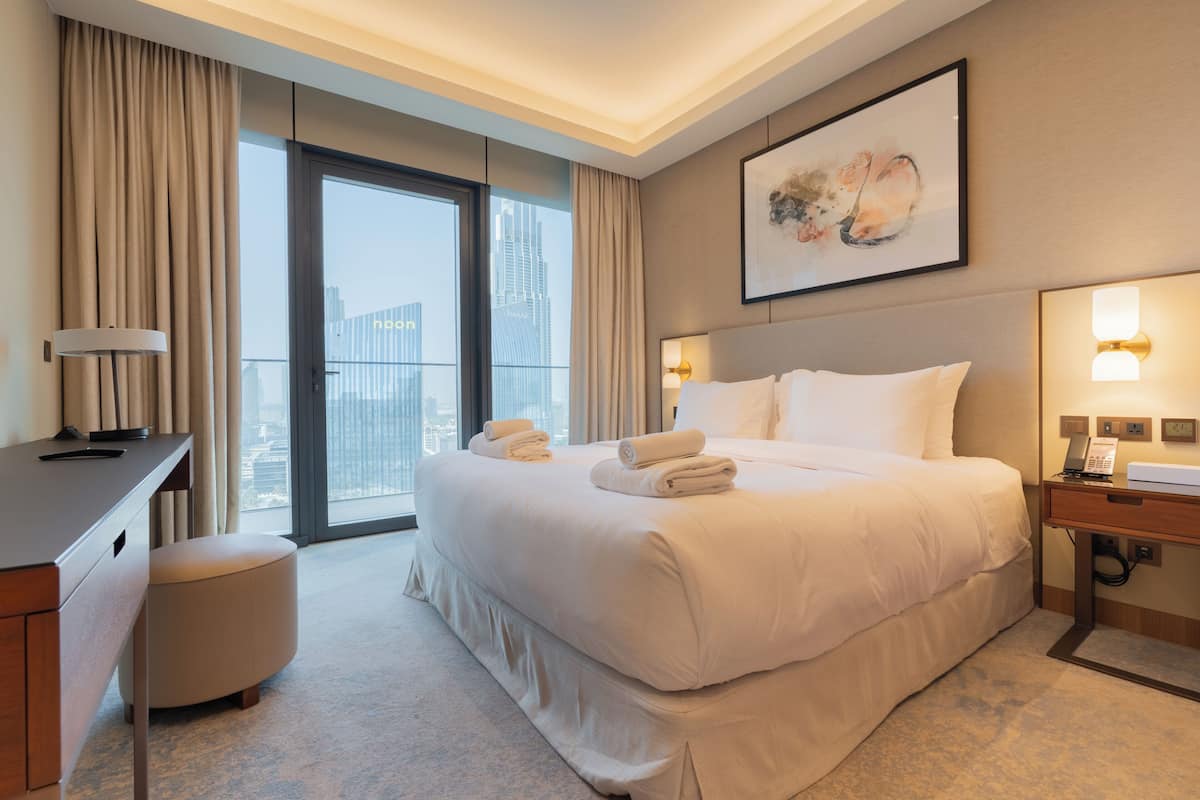 A hotel room with a large bed, a desk, a chair, a bedside table, and a window with a city view.
