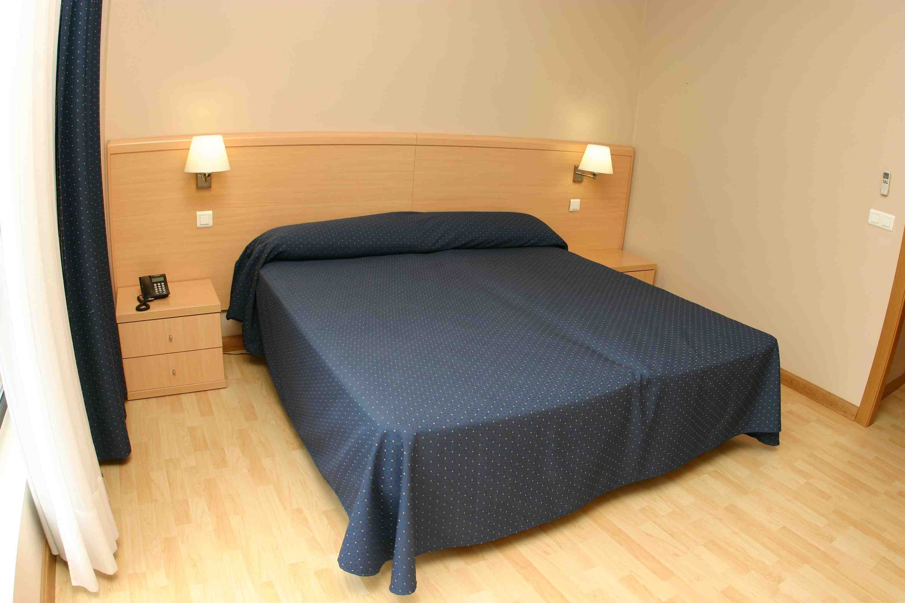 A neatly made bed with a dark blue bedspread, two bedside lamps, a wooden headboard, a small bedside table with a phone, and a wooden floor.