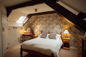 Un dormitorio con techo inclinado, una cama con ropa de cama blanca, dos lámparas de noche, una mesita de noche de madera, un aparador y un papel tapiz con motivos florales.