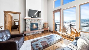 Un salon spacieux doté d’une cheminée en pierre, de meubles en cuir et de grandes fenêtres offrant une vue sur les montagnes.