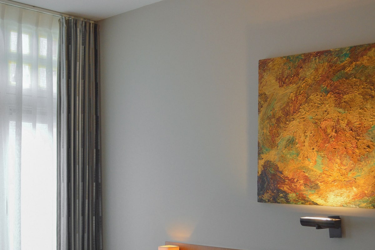 A hotel room with a bed, a nightstand, a wall-mounted lamp, a window with curtains, and a painting on the wall.