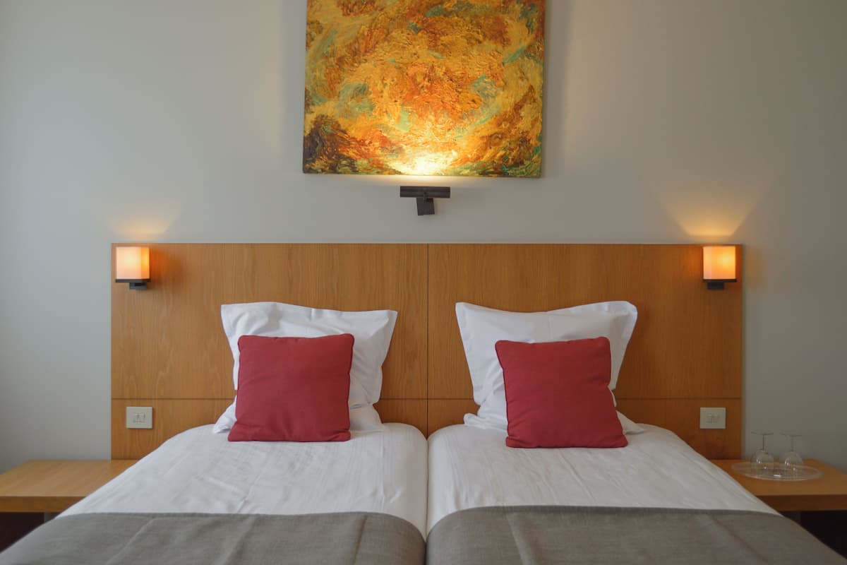 A hotel room with two beds, each with white bedding and red pillows, a wooden headboard, and a large abstract painting above the beds.