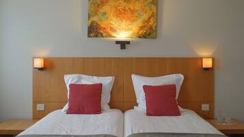 Une chambre d’hôtel avec deux lits, chacun agrémenté de draps blancs et d’oreillers rouges, d’une tête de lit en bois et d’un grand tableau abstrait au-dessus des lits.