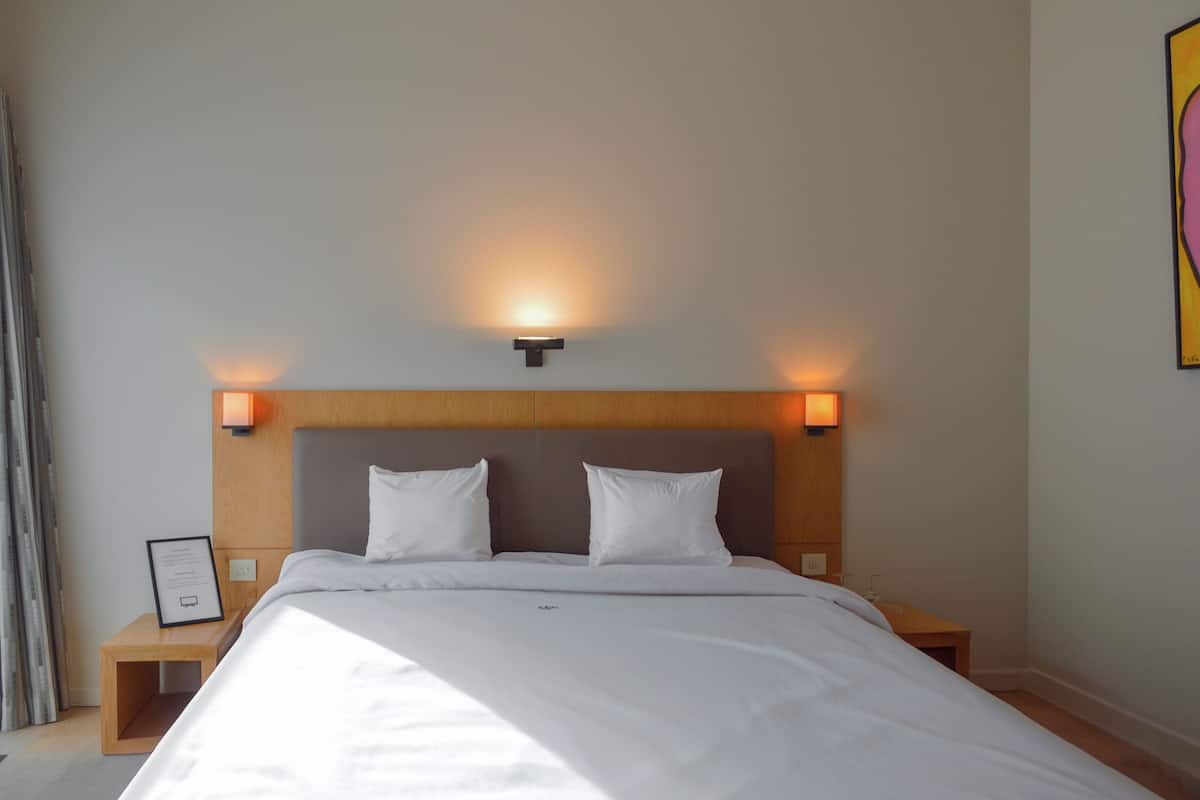 A neatly made bed with white linens and two pillows, a wooden headboard, and two bedside lamps.