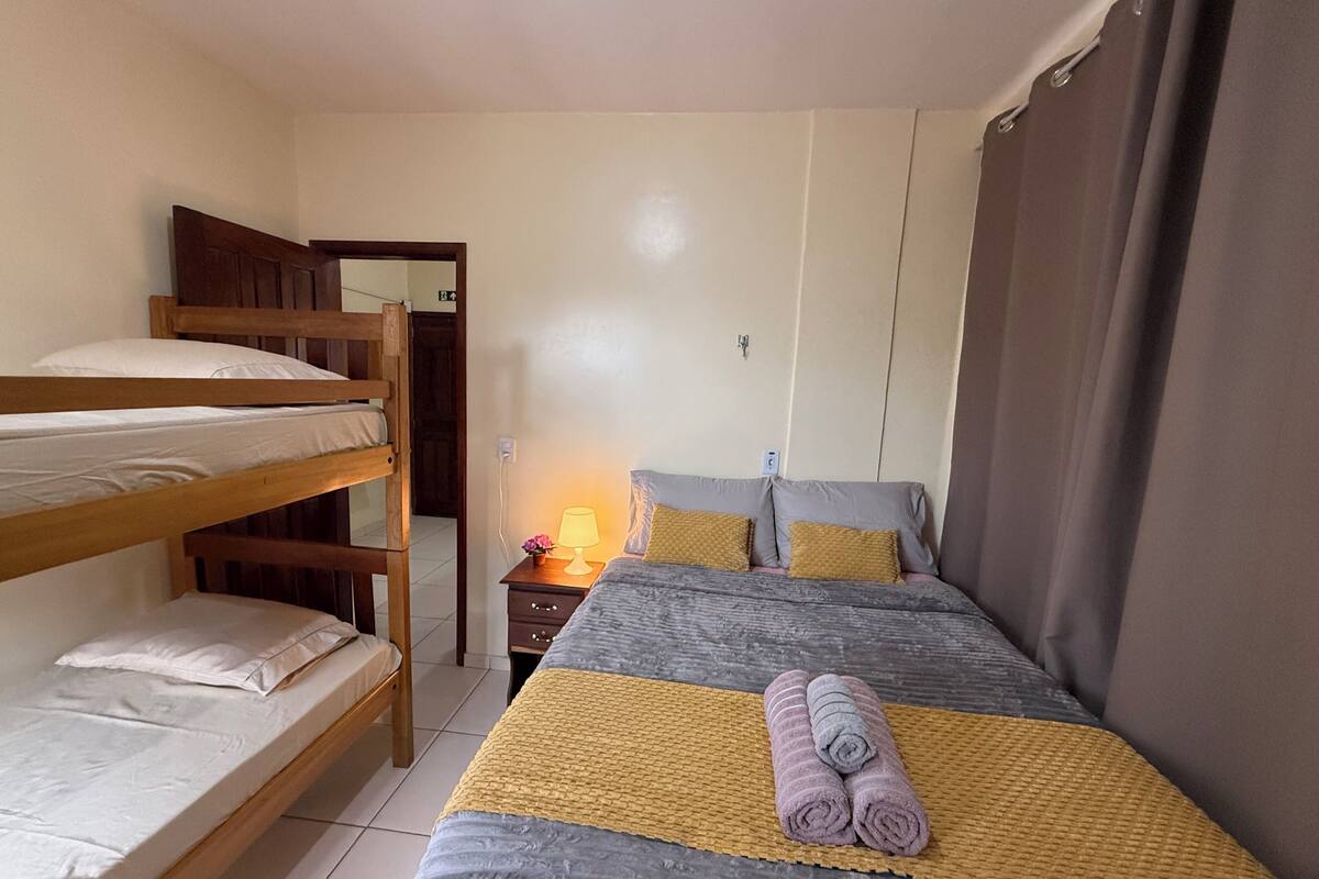 A dormitory room with bunk beds, a single bed, a bedside table with a lamp, and a window with curtains.