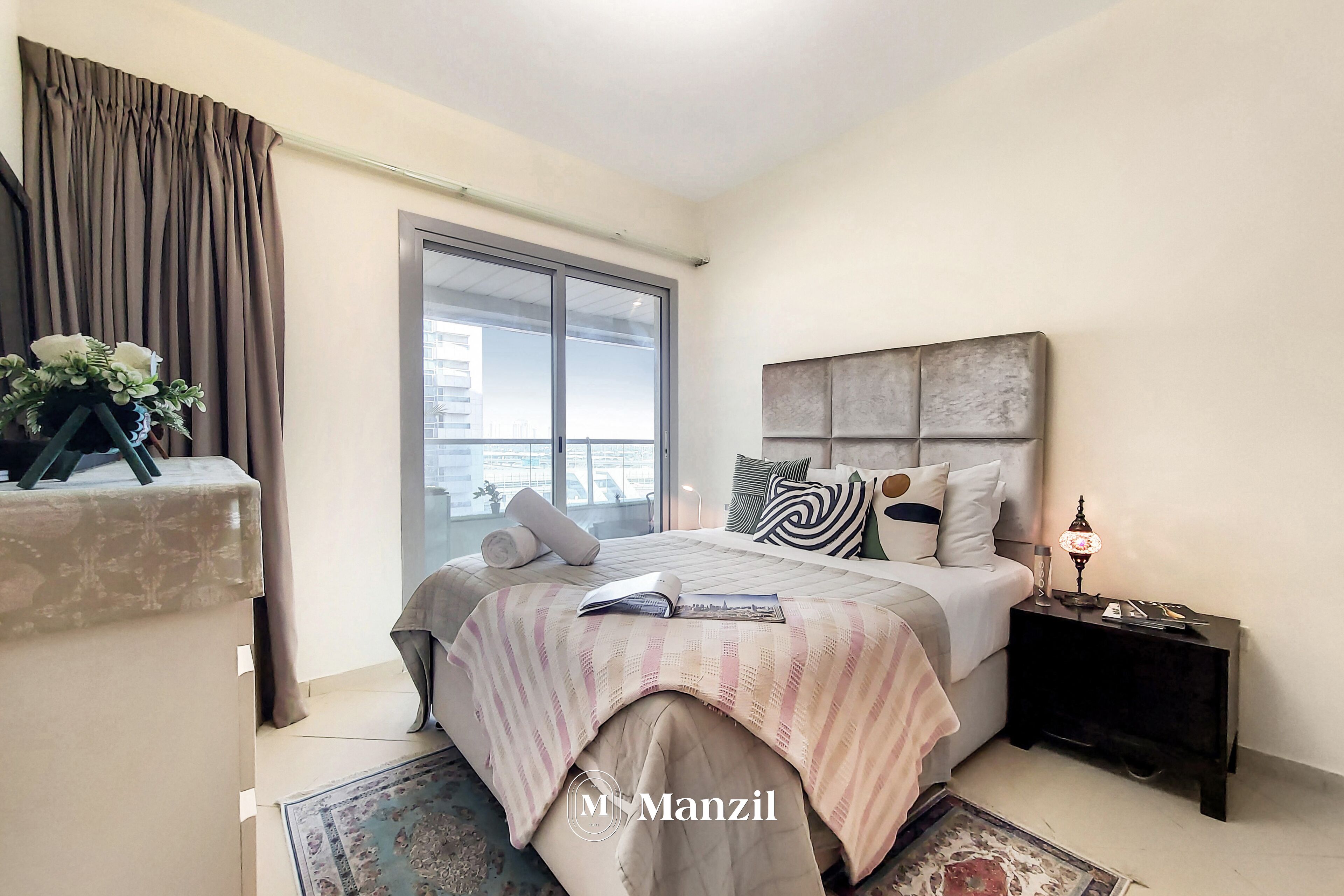 A bedroom with a large window, a bed with a striped bedspread, a bedside table with a lamp, and a decorative vase on a shelf.