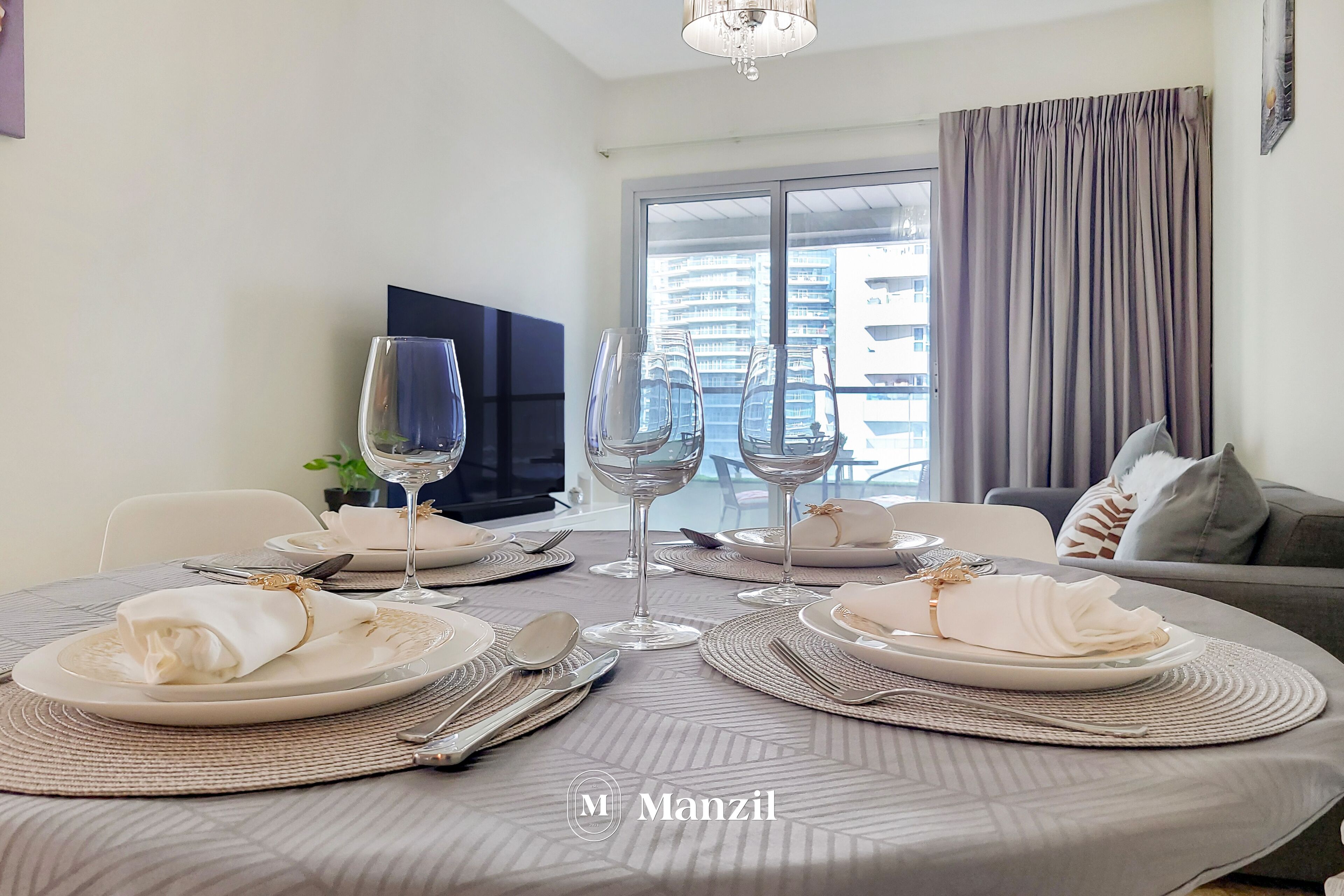 Un coin repas avec une table ronde dressée pour quatre personnes, agrémentée d’assiettes blanches, de couverts en argent et de verres à vin. Un téléviseur est fixé au mur, et une fenêtre avec des rideaux est visible.