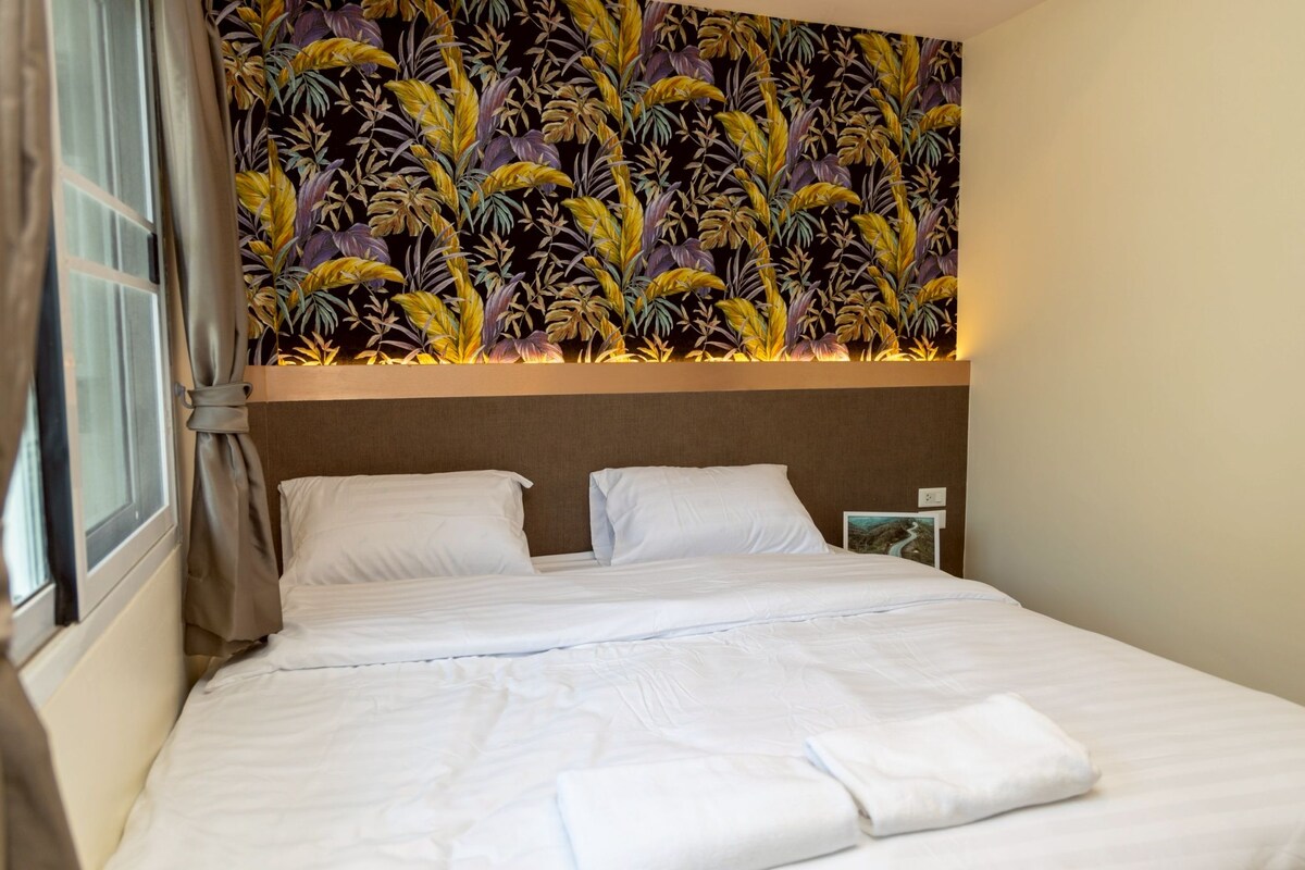 A hotel room with a bed, curtains, and a wall with a tropical leaf pattern.
