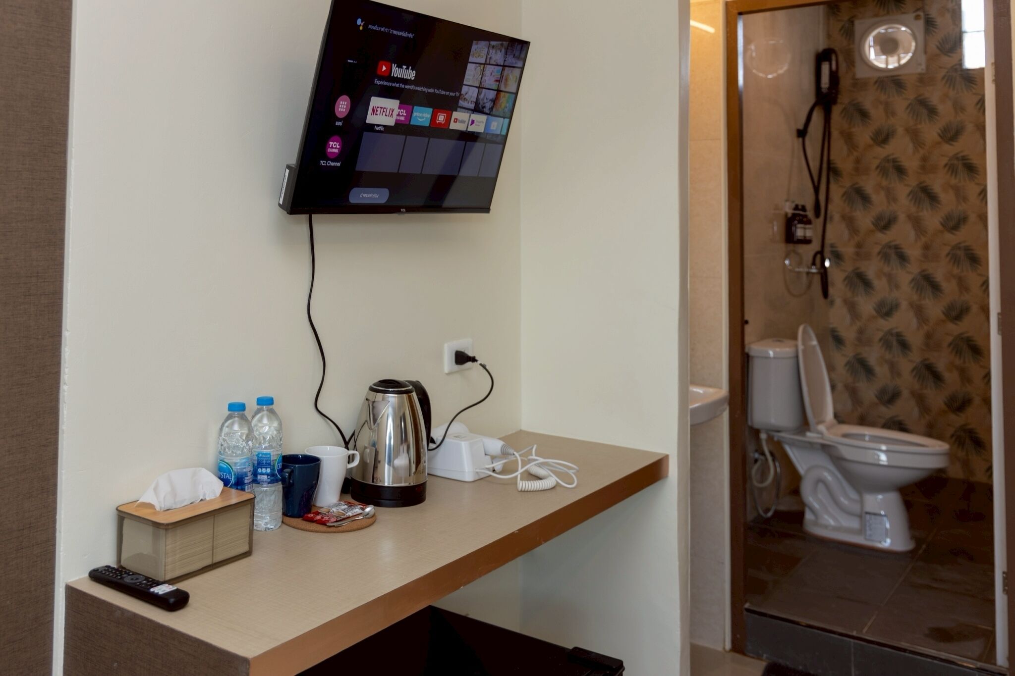 A hotel room with a mounted TV displaying streaming services, a coffee maker, bottled water, a remote control, and a toilet with a shower.